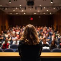 Stock image show back of woman facing an audience as if ready to deliver an address.